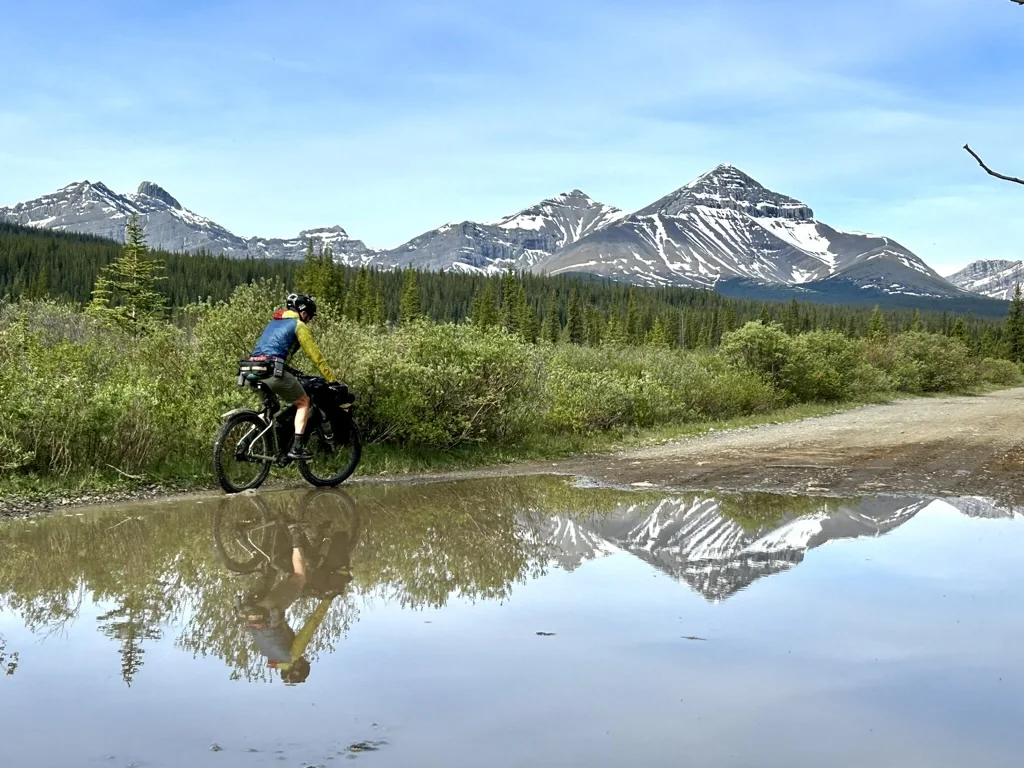 Bikepacker riding through a wet Alberta backroad with mountain reflections